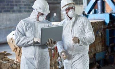 Portrait of two workers wearing biohazard suits using laptop  in industrial warehouse of modern waste processing plant against recyclable cardboard in background