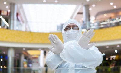 man in coverall disposable anti-epidemic antibacterial isolation suit shows a definitive stopping gesture with two hands against the background of some interior of public space